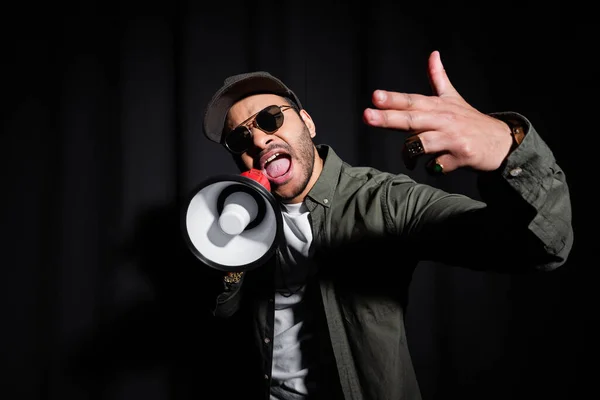 Émotionnel Moyen-Orient hip hop interprète dans les lunettes de soleil et casquette chantant et tenant haut-parleur sur noir — Photo de stock