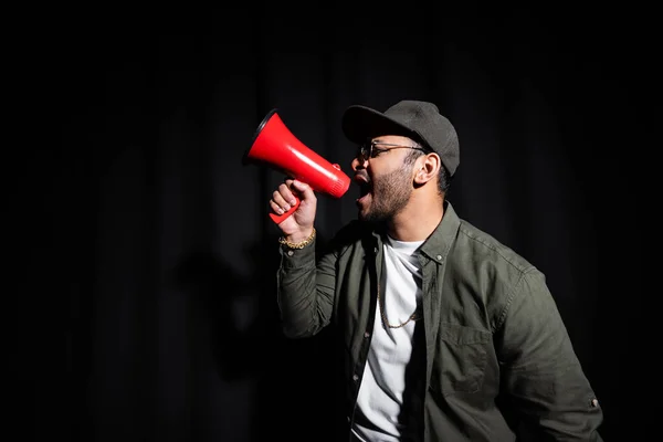 Vue latérale de l'artiste hip hop émotionnel du Moyen-Orient en lunettes de soleil et casquette chantant et tenant haut-parleur sur noir — Photo de stock
