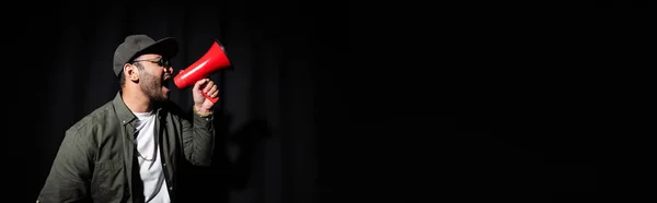 Вид сбоку эмоционального исполнителя хип-хопа Среднего Востока в солнцезащитных очках и кепке, поющего и держащего громкоговоритель на черном баннере — стоковое фото