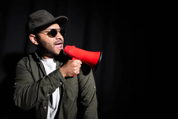 Émotionnel indien hip hop interprète dans des lunettes de soleil et casquette chantant et tenant haut-parleur sur noir — Photo de stock