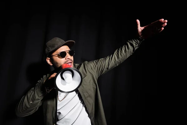 Эмоциональный ближневосточный хип-хоп исполнитель в солнцезащитных очках и кепке разговаривающий в громкоговорителе на черном — стоковое фото