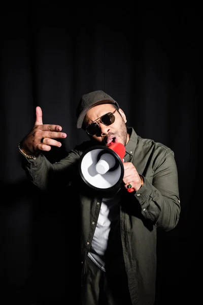 Émotionnel Moyen-Orient hip hop interprète en lunettes de soleil et casquette parler en mégaphone sur noir — Photo de stock