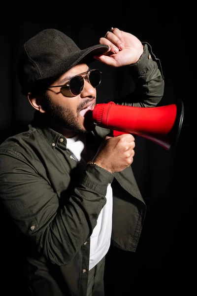 Cantante hip hop indiano in occhiali da sole e cappuccio urlando in megafono su nero — Foto stock