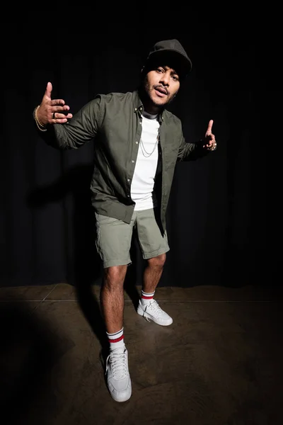 Full length of stylish indian hip hop singer in cap gesturing while standing on black — Stockfoto