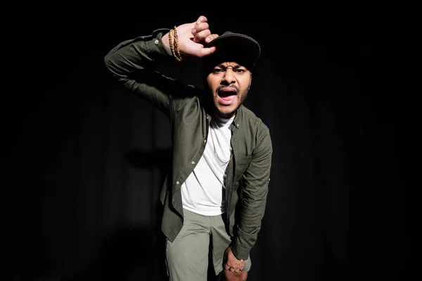 Emotional and stylish indian hip hop singer adjusting cap while singing on black — Fotografia de Stock