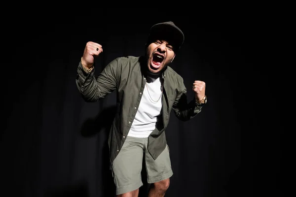 Emocional y elegante cantante indio de hip hop en gorra cantando en voz alta en negro - foto de stock