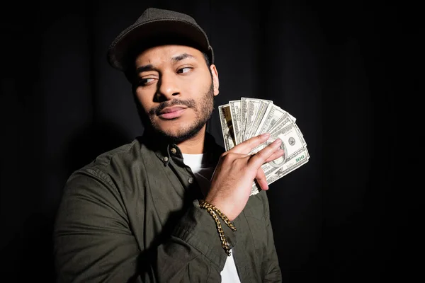Stylish indian hip hop singer in cap holding dollar banknotes isolated on black — Fotografia de Stock