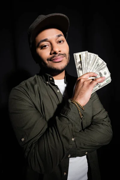 Élégant chanteur hip hop oriental en bonnet tenant des billets en dollars isolés sur noir — Photo de stock