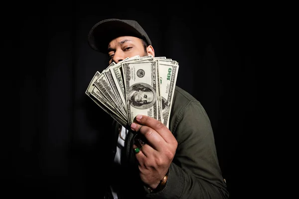 Bearded indian hip hop performer in cap covering face with dollar banknotes on black - foto de stock