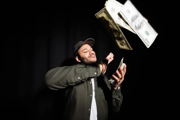 Rich indian hip hop performer in cap throwing dollar banknotes on black - foto de stock