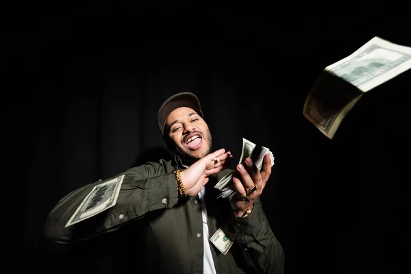Excited and rich indian hip hop performer in cap throwing dollar banknotes on black - foto de stock