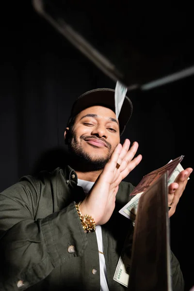 Pleased indian hip hop performer in cap throwing dollar banknotes on black — Fotografia de Stock