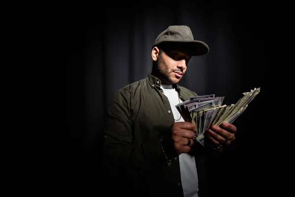 Rich indian hip hop performer in cap counting dollar banknotes on black - foto de stock