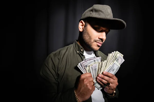 Rich indian hip hop performer in cap holding dollar banknotes on black — Fotografia de Stock