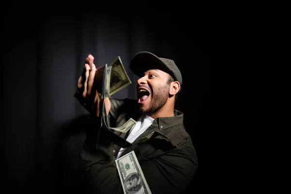 Stupito artista hip hop indiano in cap gettando banconote in dollari sul nero — Foto stock