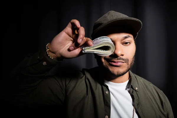 Rich indian hip hop performer in cap covering eye with bundle of dollar banknotes on black — Foto stock
