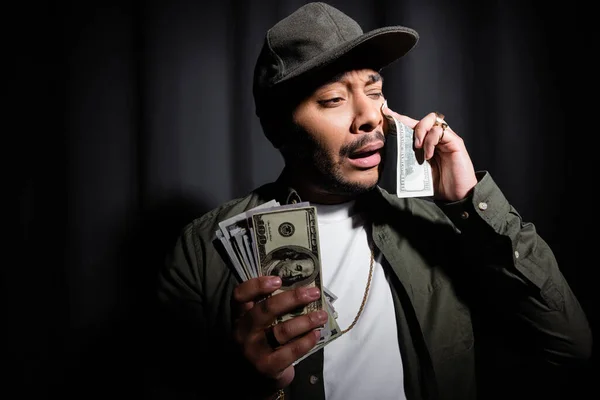 Triste artista indio de hip hop en gorra llorando y limpiando lágrima con billete de dólar en negro - foto de stock