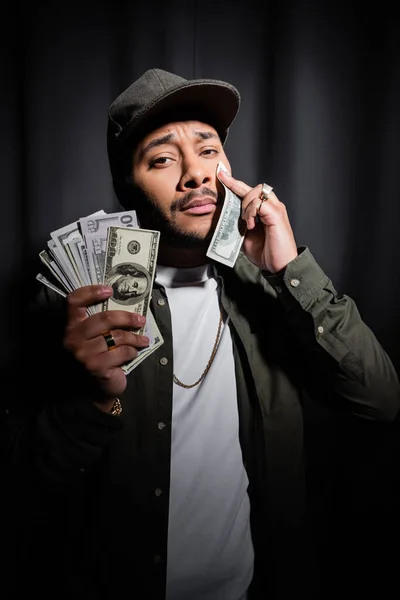 Sad indian hip hop performer in cap wiping tear with dollar banknote on black — Fotografia de Stock