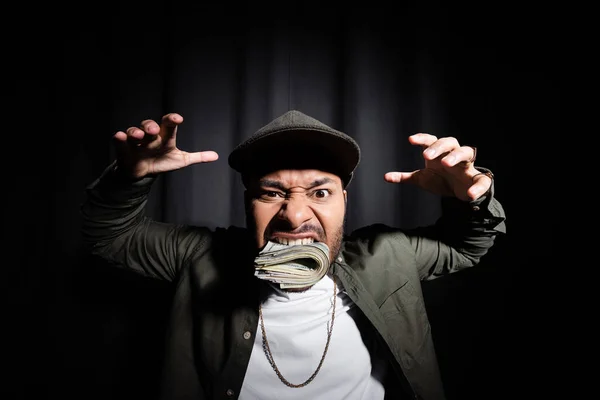Rich indian hip hop performer in cap biting bundle of dollar banknotes and showing spooky gesture on black — Foto stock