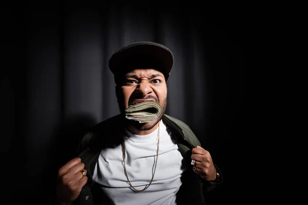 Indian hip hop performer in cap biting bundle of dollar banknotes on black — Stock Photo