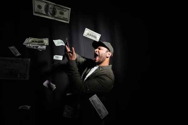 Excited eastern hip hop performer in cap throwing dollar banknotes on black — Photo de stock