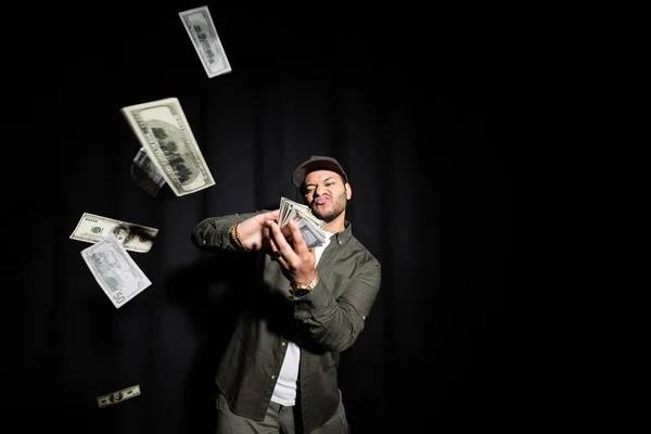 Rich indian hip hop performer in cap throwing blurred dollar banknotes on black — Stock Photo