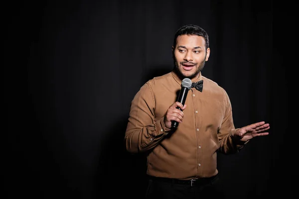 Indian stand up comedian with bow tie holding microphone during monologue on black — Foto stock