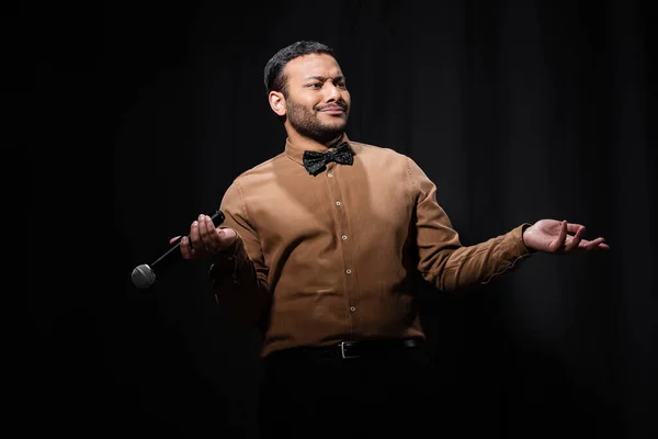 Skeptical indian comedian in shirt and bow tie gesturing while holding microphone during monologue on black - foto de stock