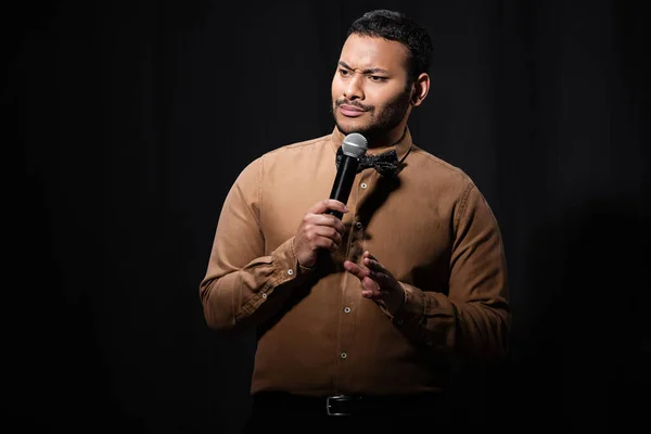Skeptical indian comedian in shirt and bow tie holding microphone during monologue on black — Fotografia de Stock