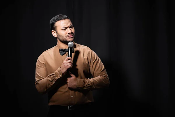 Comediante indio ofendido en camisa y pajarita sosteniendo micrófono durante monólogo en negro - foto de stock