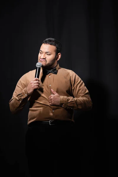 Indischer Stand-up-Comedian grimmig und mit erhobenem Daumen beim Erzählen von Witzen ins Mikrofon auf schwarz — Stockfoto
