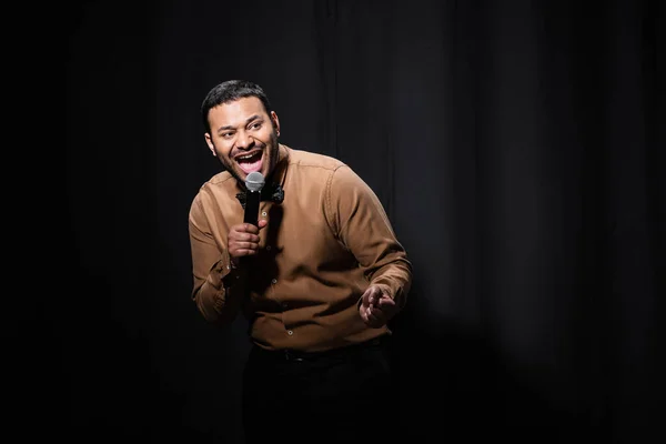 Amazed indian stand up comedian telling jokes into microphone on dark stage on black — Stock Photo