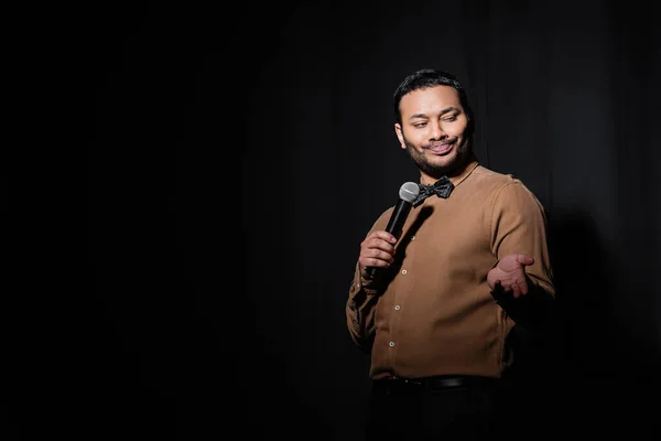 Skeptical indian stand up comedian telling joke into microphone on dark stage on black — Fotografia de Stock