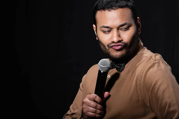 Skeptical indian stand up comedian telling joke into microphone and pouting lips isolated on black — Fotografia de Stock