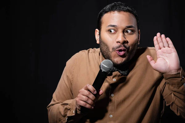 Emotional indian stand up comedian showing stop gesture while telling jokes into microphone isolated on black - foto de stock