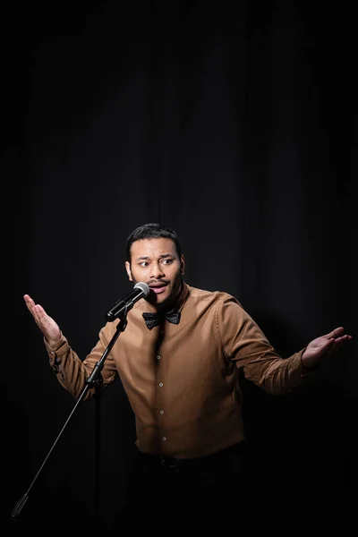 Indiano stand up comico gesticolando mentre racconta barzellette in microfono su stand isolato su nero — Foto stock