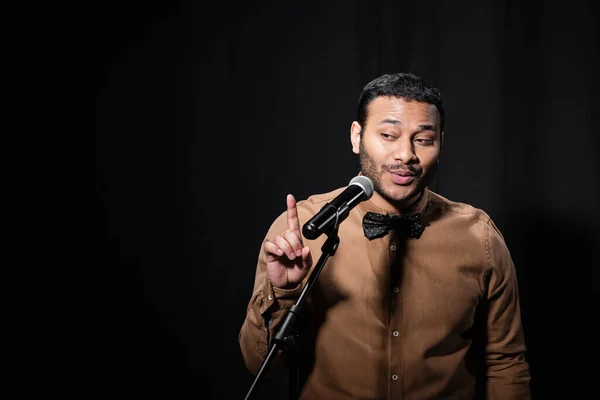 Indian stand up comedian pointing with finger while telling jokes into microphone on stand on black background - foto de stock