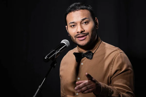 Indian stand up comedian telling jokes into microphone and pointing with finger while looking at camera isolated on black — Stock Photo