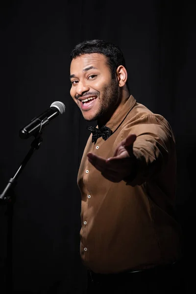 Amazed indian stand up comedian telling jokes into microphone on stand isolated on black — Stock Photo