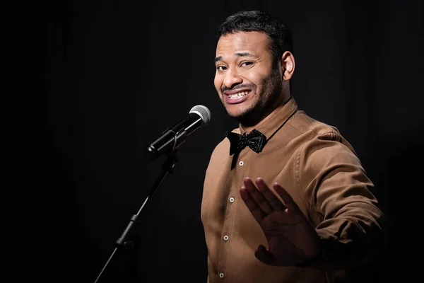 Displeased indian stand up comedian showing stop gesture while telling jokes into microphone isolated on black — Photo de stock