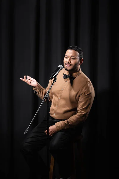 Comico indiano esibendosi stand up comedy e raccontare barzellette in microfono mentre seduto su nero — Foto stock