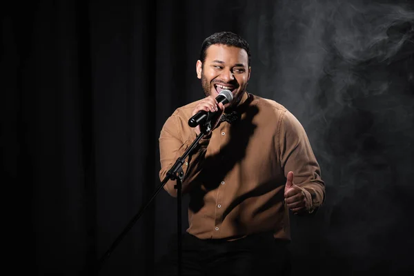 Fröhlicher indischer Komiker sitzt auf Stuhl und führt Stand-up-Comedy in Mikrofon auf schwarz mit Rauch — Stockfoto