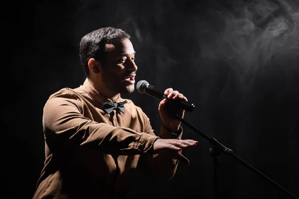 Indian comedian performing stand up comedy into microphone and looking away on black with smoke - foto de stock