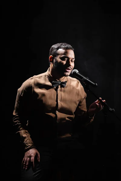 Indian stand up comedian telling jokes into microphone on dark stage isolated on black — Stock Photo