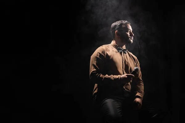 Comediante indio sosteniendo micrófono y mirando hacia otro lado en negro con humo - foto de stock