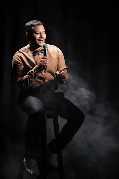 Indian comedian sitting and performing stand up comedy into microphone on black with smoke — Foto stock