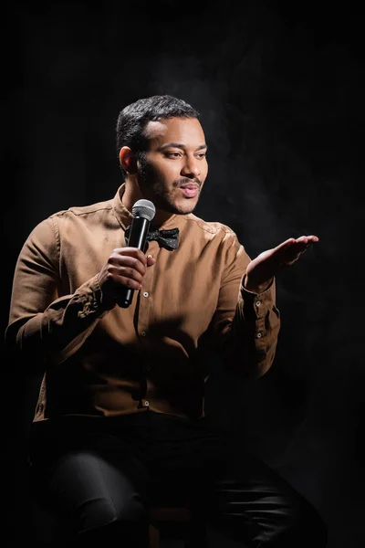 Indian comedian sitting and pouting lips while performing stand up comedy into microphone on black - foto de stock