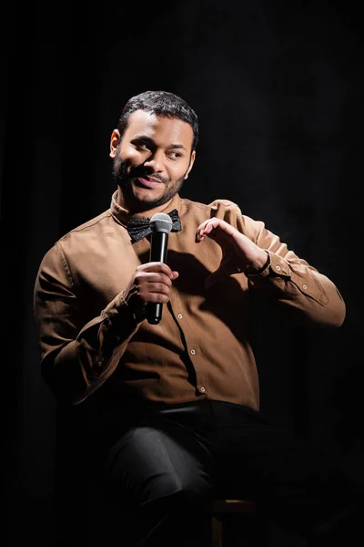 Улыбающийся индийский комик, сидящий и исполняющий стенд-ап комедию на черном — стоковое фото