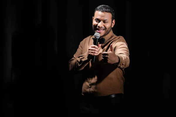 Joyeux comédien moyen-oriental montrant comme et performant stand up comédie dans microphone isolé sur noir — Photo de stock