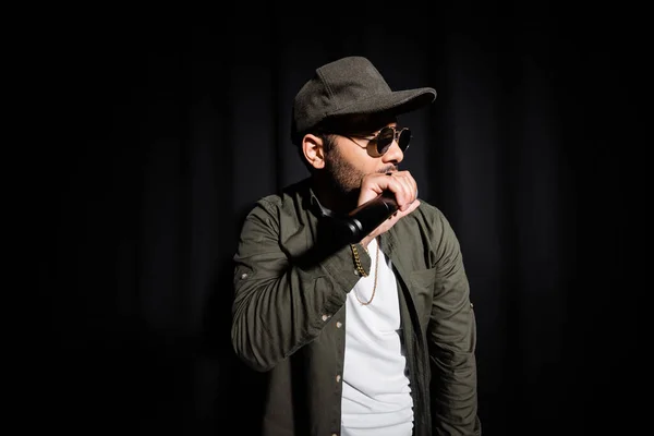 Middle east hip hop singer in cap and sunglasses singing into microphone on black - foto de stock